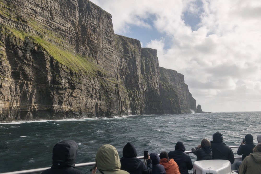 A cliff-cruise boat directly beneath the Cliffs of Moher, passengers at the rail looking up at the vast layered rock face streaked green above the grey Atlantic swell.