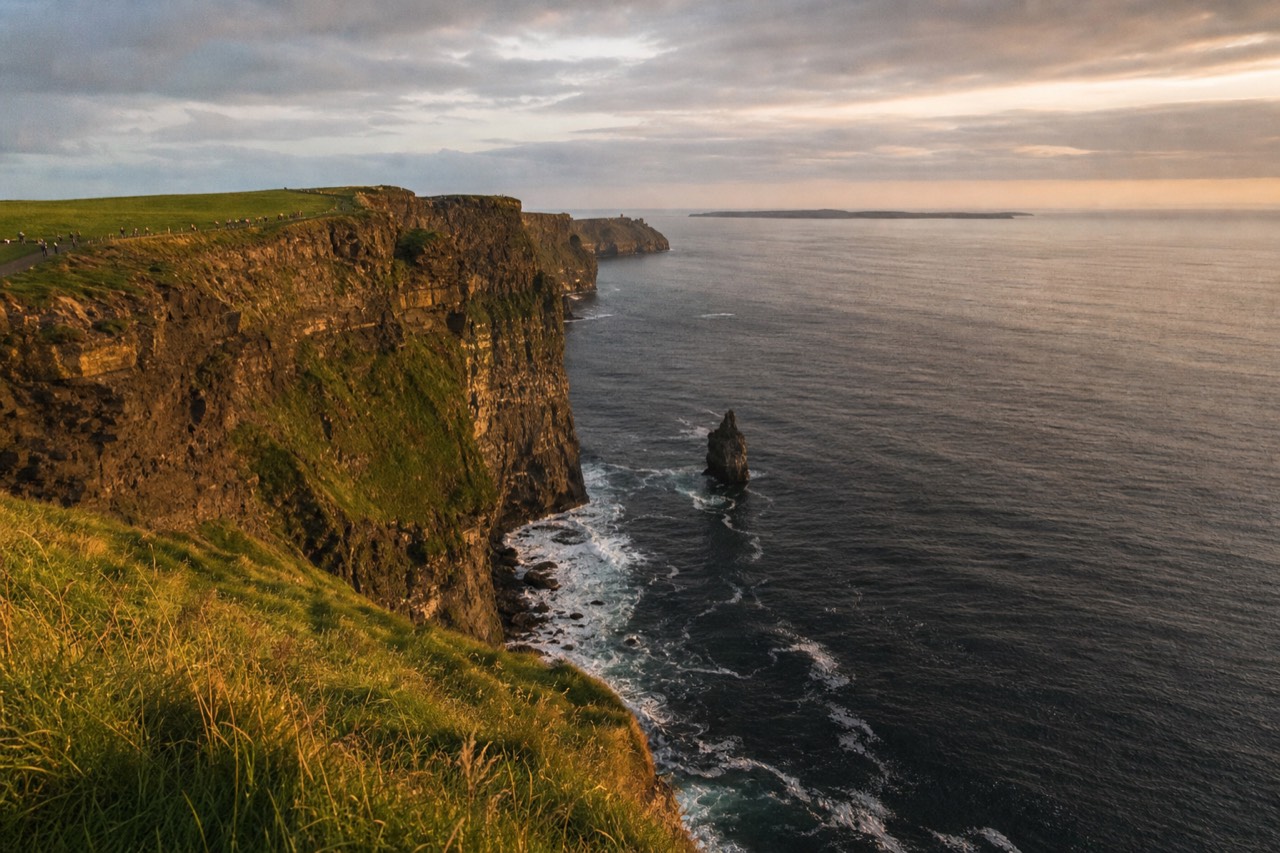 The Cliffs of Moher rising from the mainland Clare coast, green farmland running to the clifftop and the low green shapes of the Aran Islands sitting out in the sea beyond.