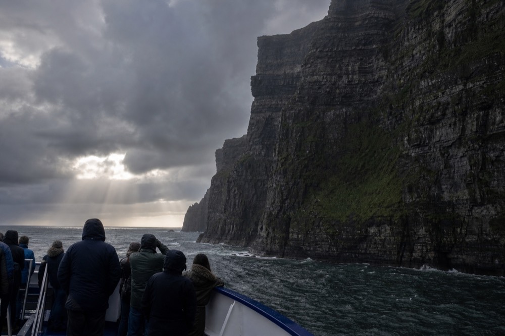 The layered rock face of the Cliffs of Moher seen from the deck of a cliff cruise, the wall rising far out of frame above passengers looking up from the rail.