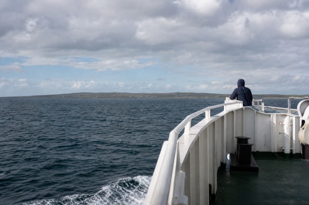 An Aran Islands ferry crossing open grey-green Atlantic water off the Galway coast, the low green line of Inis Mór on the horizon under a broad cloudy sky.