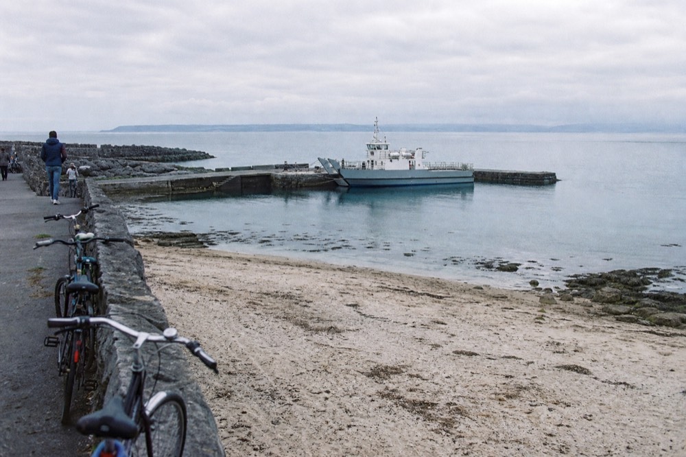 The small stone harbour of Inis Oírr with a sandy beach and bikes leaning against a low wall, a ferry moored alongside and the Clare coast faint across the water.