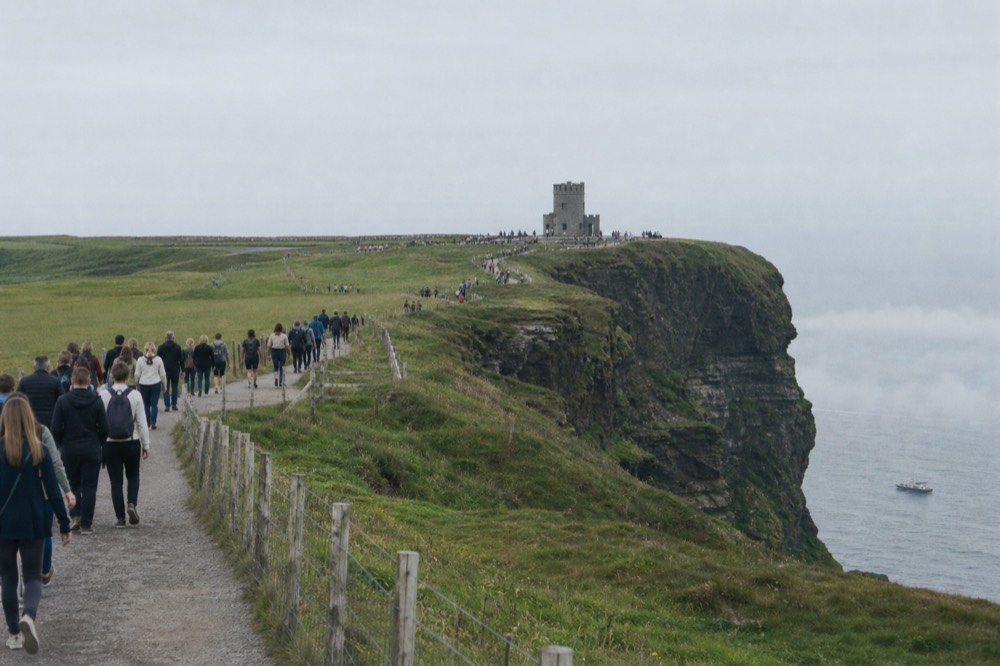 Summer crowds along the clifftop path at the Cliffs of Moher under hazy sunshine, O'Brien's Tower on the headland and the Atlantic pale behind a thin band of sea fog.