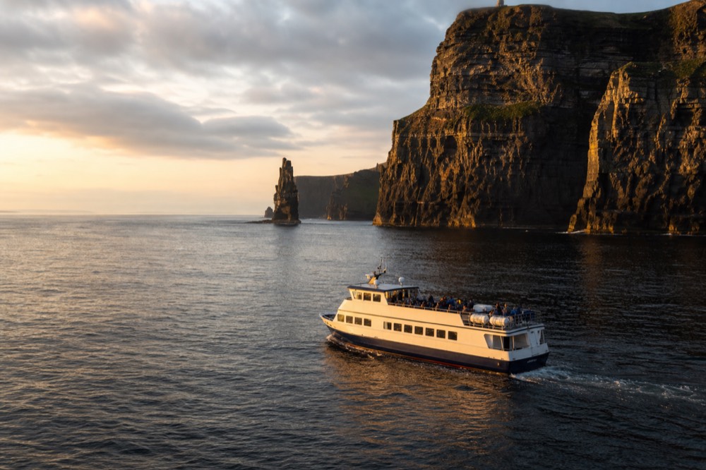 A quiet September afternoon at the base of the Cliffs of Moher, a cruise boat passing beneath the towering layered rock face with soft golden light on the sea.
