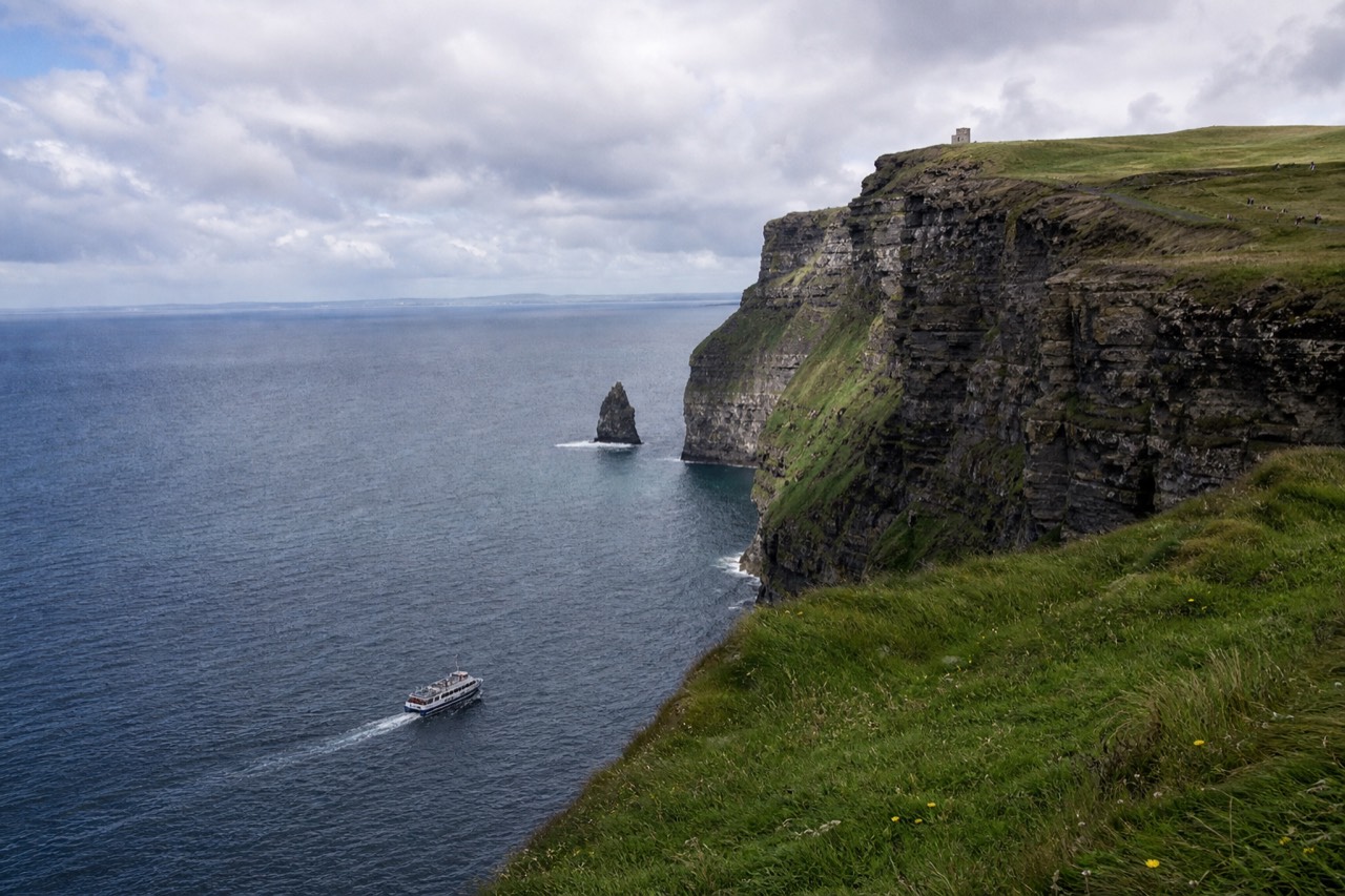 The Cliffs of Moher on a bright late-spring morning, green clifftops above a calm blue Atlantic, a small cruise boat tracking the base of the rock wall far below.