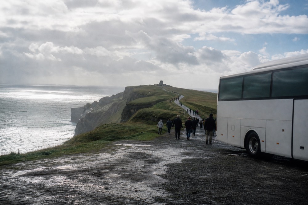 A tour coach parked near the green clifftop above the Cliffs of Moher, a group of visitors walking the path toward O'Brien's Tower under a broad Atlantic sky.