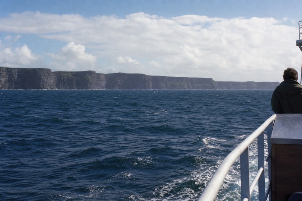 The Cliffs of Moher seen at a distance from an Aran Islands ferry on the return leg, the dark cliff line stretching along the horizon above choppy Atlantic swell.