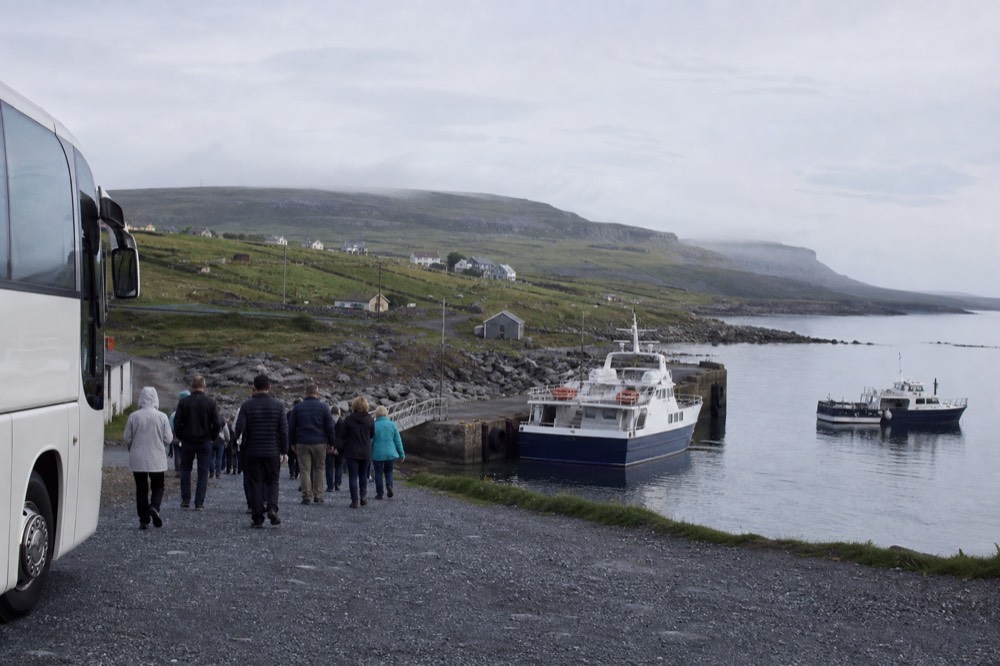 A tour coach parked near Doolin village with green Burren hills behind, passengers walking down toward the pier where the cliff cruise boats wait.
