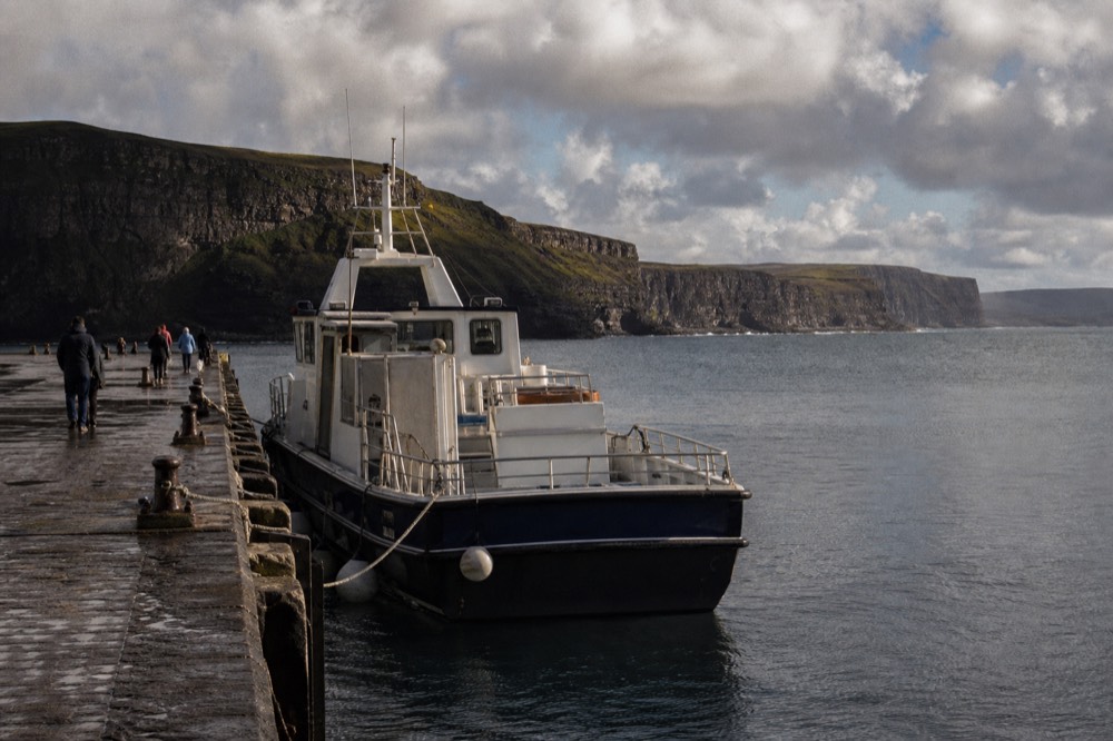 Doolin Pier on a calm morning, a small cliff cruise boat tied alongside the stone quay with the flat Atlantic and the low Clare coastline beyond.