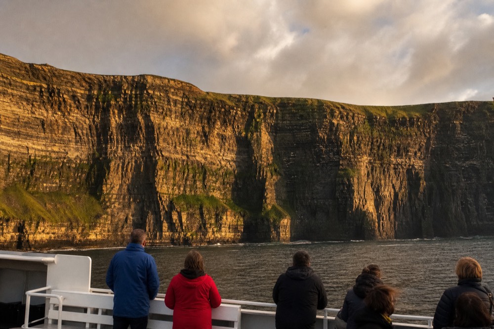 The full face of the Cliffs of Moher seen from a boat at sea level, horizontal dark rock bands streaked with green rising far above a thin line of white surf.