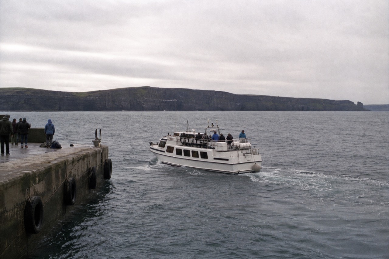 A white passenger boat leaving the stone jetty at Doolin Pier under a wide grey sky, the Cliffs of Moher rising as a dark line to the south.