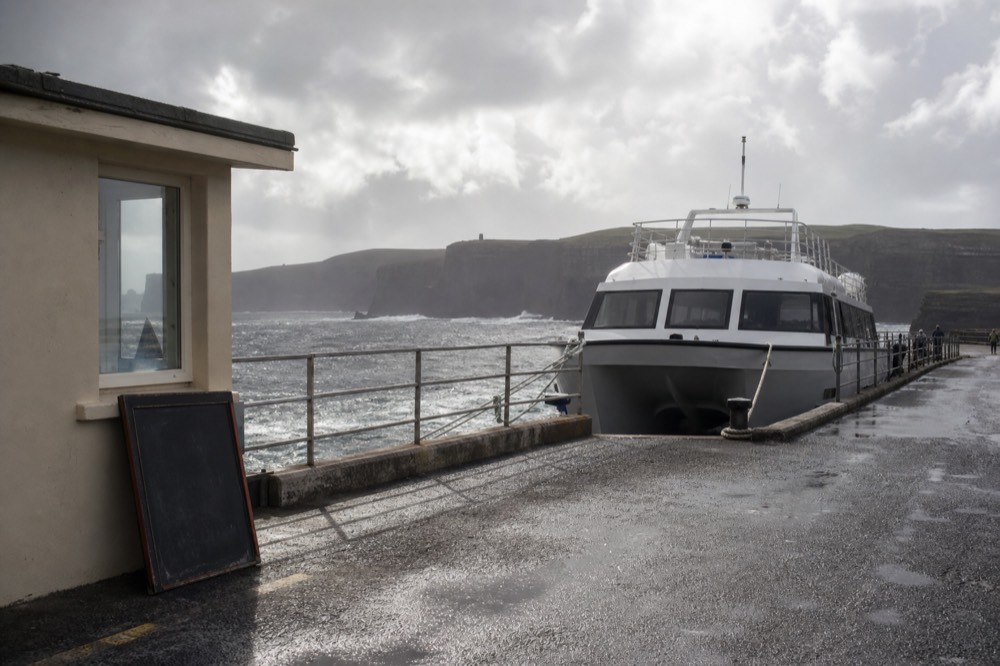 A grey, windy day at Doolin Pier with the sea choppy and a cliff cruise boat tied up, a chalkboard sign noting a weather cancellation by the ticket hut.