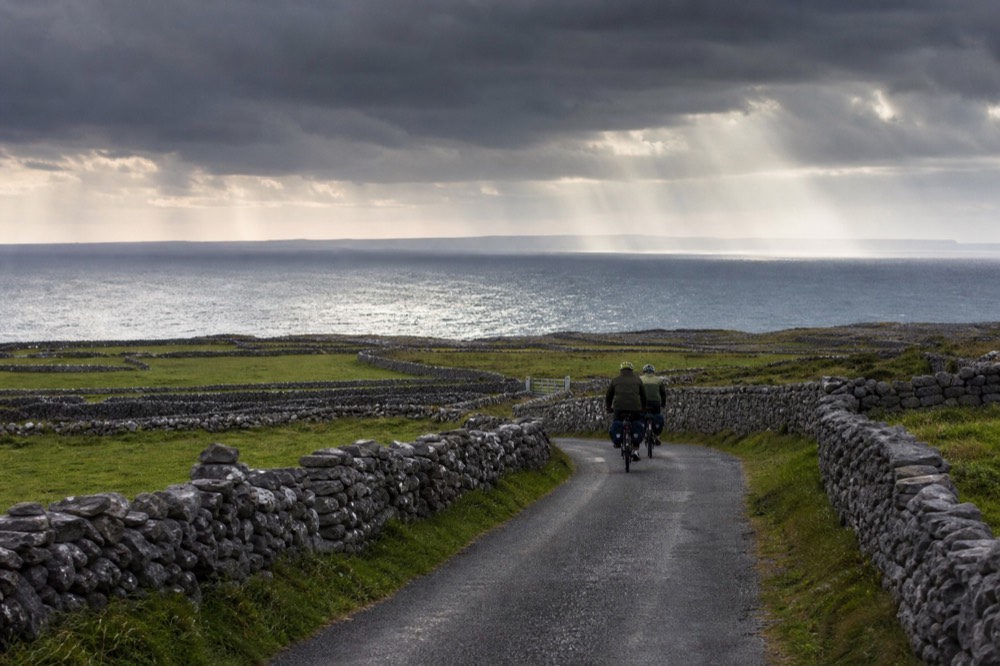 Cyclists on a quiet lane on an Aran island, low stone walls and green fields around them, the Atlantic and a hazy mainland coastline in the distance.