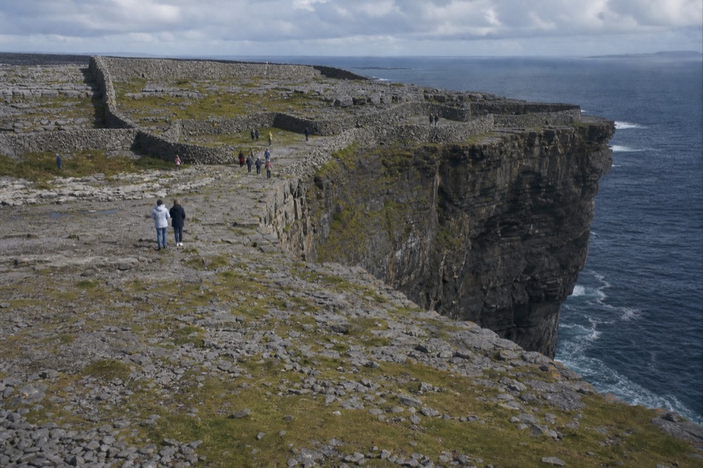 The stone ramparts of Dún Aonghasa on Inis Mór perched at the edge of high sea cliffs, the Atlantic far below and a scatter of visitors near the drop.