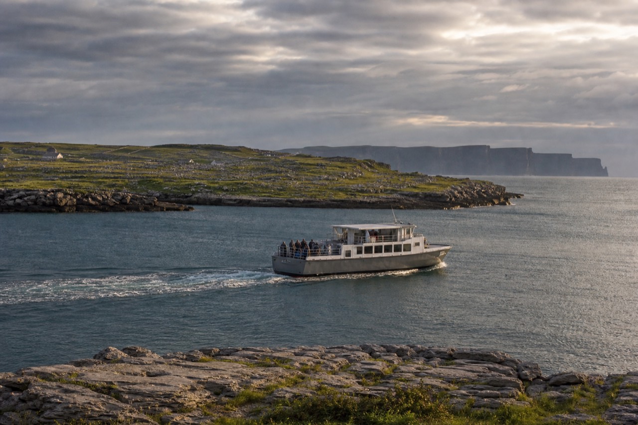 A tour boat crossing calm blue water between an Aran island and the mainland, the green ridge of Inis Oírr behind and the dark line of the Cliffs of Moher on the far horizon.