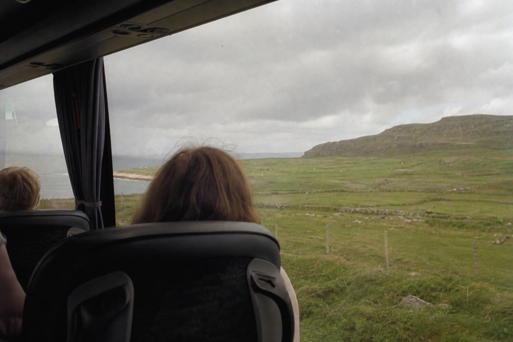The interior of a tour coach seen from a rear seat, passengers looking out at green Irish countryside sliding past the windows under a soft grey sky.