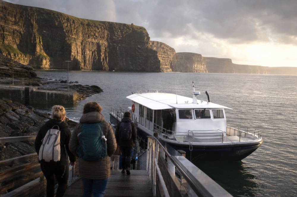 Passengers walking down the pier at Doolin toward a waiting cliff cruise boat, day bags on shoulders, the calm harbour water and the Clare coast behind them.