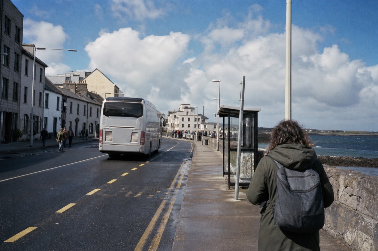 A traveller with a small backpack waiting at a Galway coach stop on a bright morning, the city's stone shopfronts behind and a tour coach pulling in.