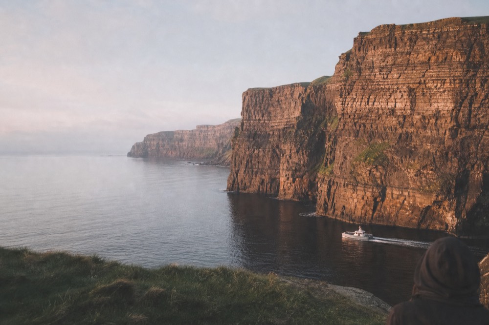 The Cliffs of Moher glowing warm orange in low evening sun, deep shadows in the rock folds, a calm Atlantic reflecting the sky as a small boat passes the base.