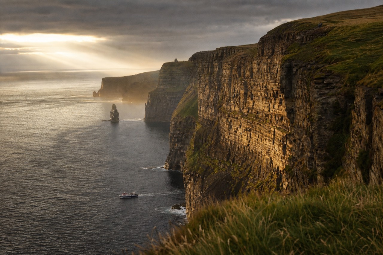 The Cliffs of Moher in warm late-afternoon light, the west-facing rock wall glowing gold above a calm sea with a small tour boat passing along the base.