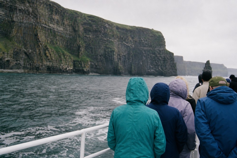 Cliff cruise passengers in waterproof jackets and hats at the boat rail on a bright but breezy May day, the layered rock face of the Cliffs of Moher rising behind them.