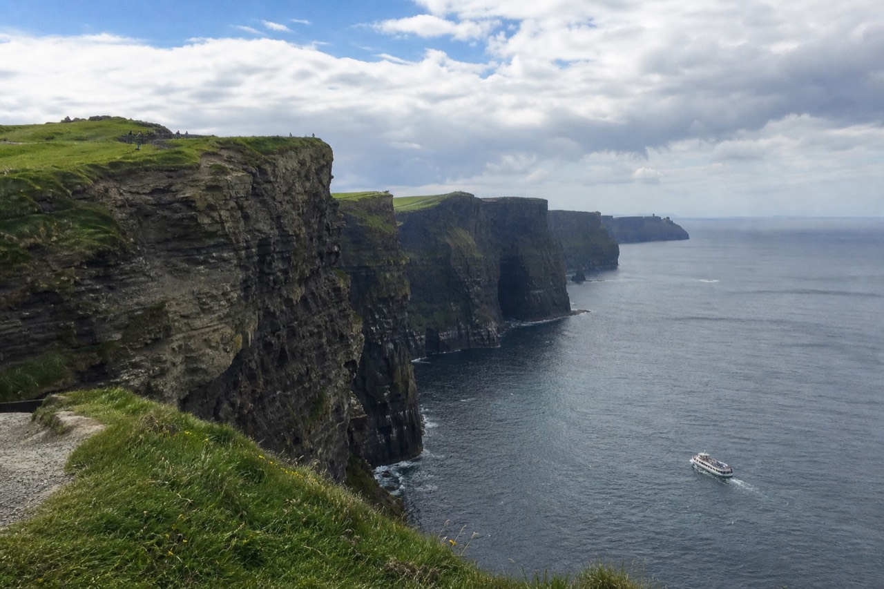 The Cliffs of Moher on a bright, breezy May morning, green grass on the clifftop, scattered white clouds over a calm blue-grey Atlantic with a small cruise boat below the rock face.