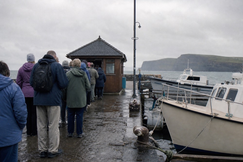 A queue of visitors at a small wooden ticket hut on Doolin Pier, boats tied up alongside the quay and a signboard listing cliff cruise departure times.