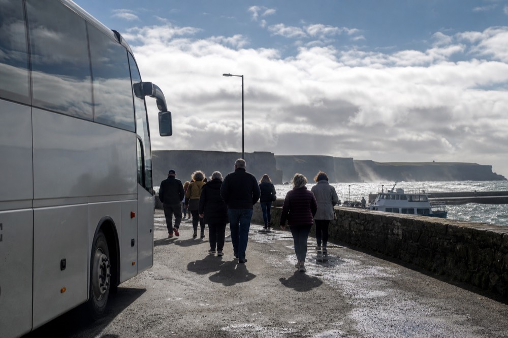 A tour coach parked near Doolin with passengers walking toward the pier, the low stone harbour wall and cliff cruise boats visible beyond.
