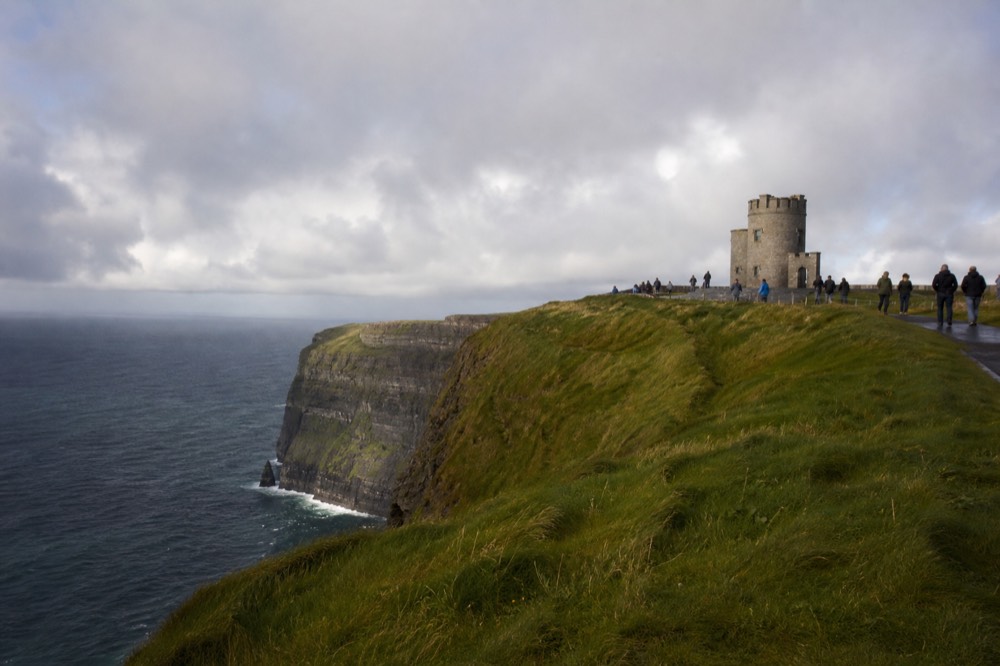 O'Brien's Tower on the highest point of the Cliffs of Moher, visitors on the flagged path beside it, the green clifftop dropping sharply to the Atlantic below.
