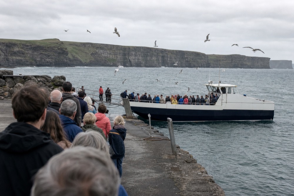 A busy Doolin Pier in summer with visitors queuing at the boarding steps, a cliff cruise boat filling with passengers and gulls wheeling over the harbour.