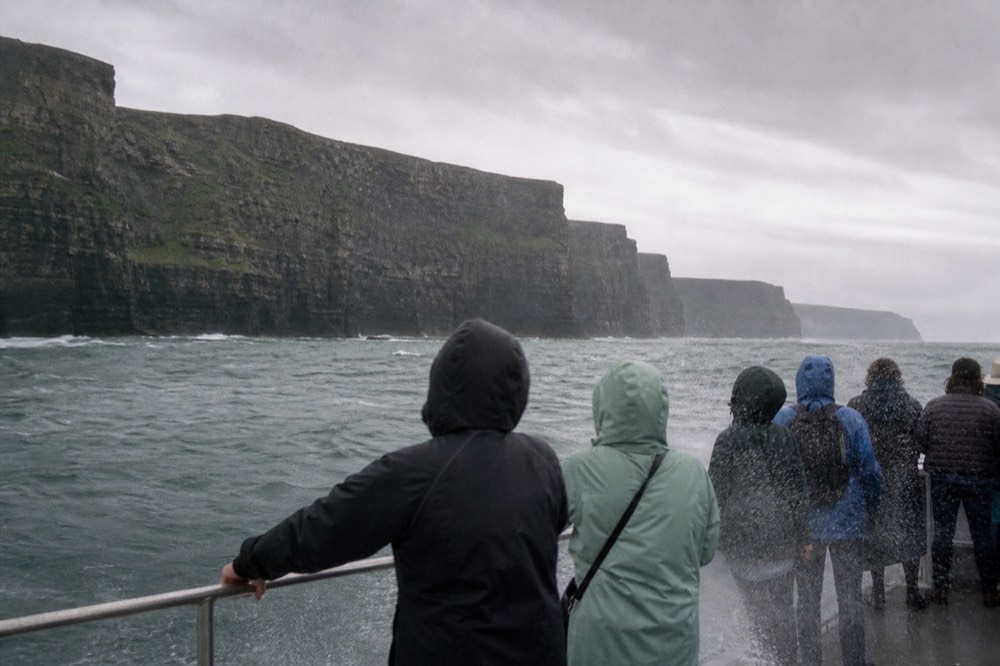 Passengers in raincoats gripping the rail of a small boat in choppy grey water below the Cliffs of Moher, spray coming over the side on a windy day.