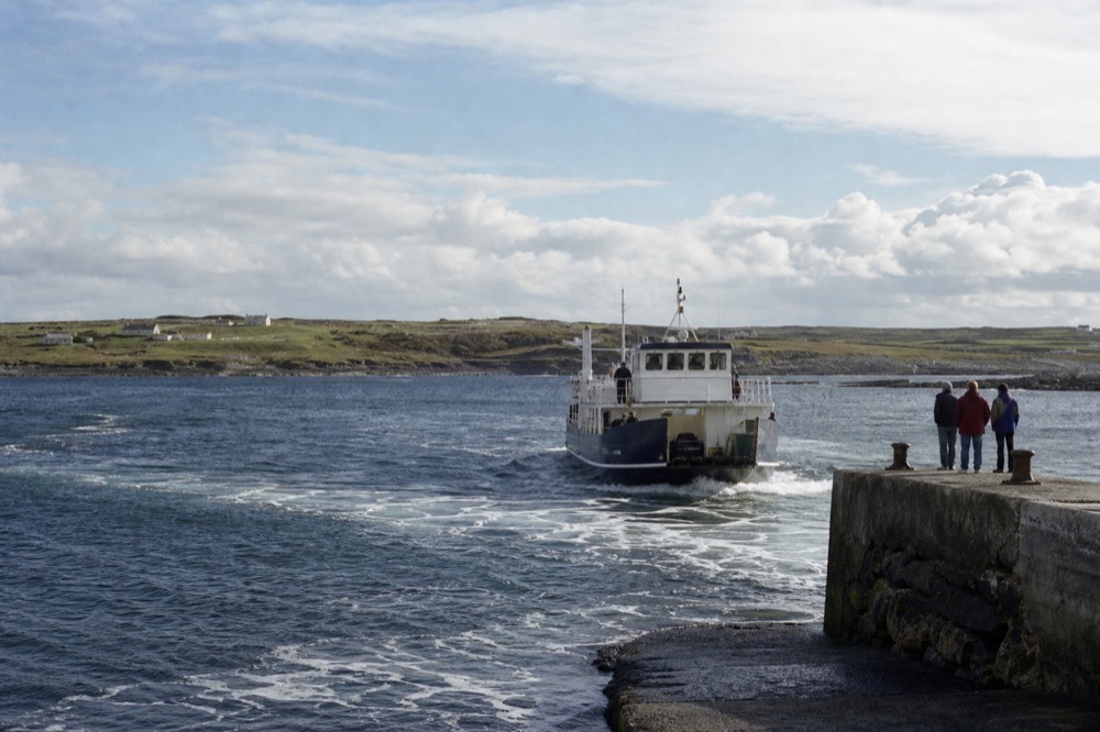 A green Aran island stretching out low over the sea, a small ferry approaching a stone harbour, the Atlantic wide and calm under a bright sky.