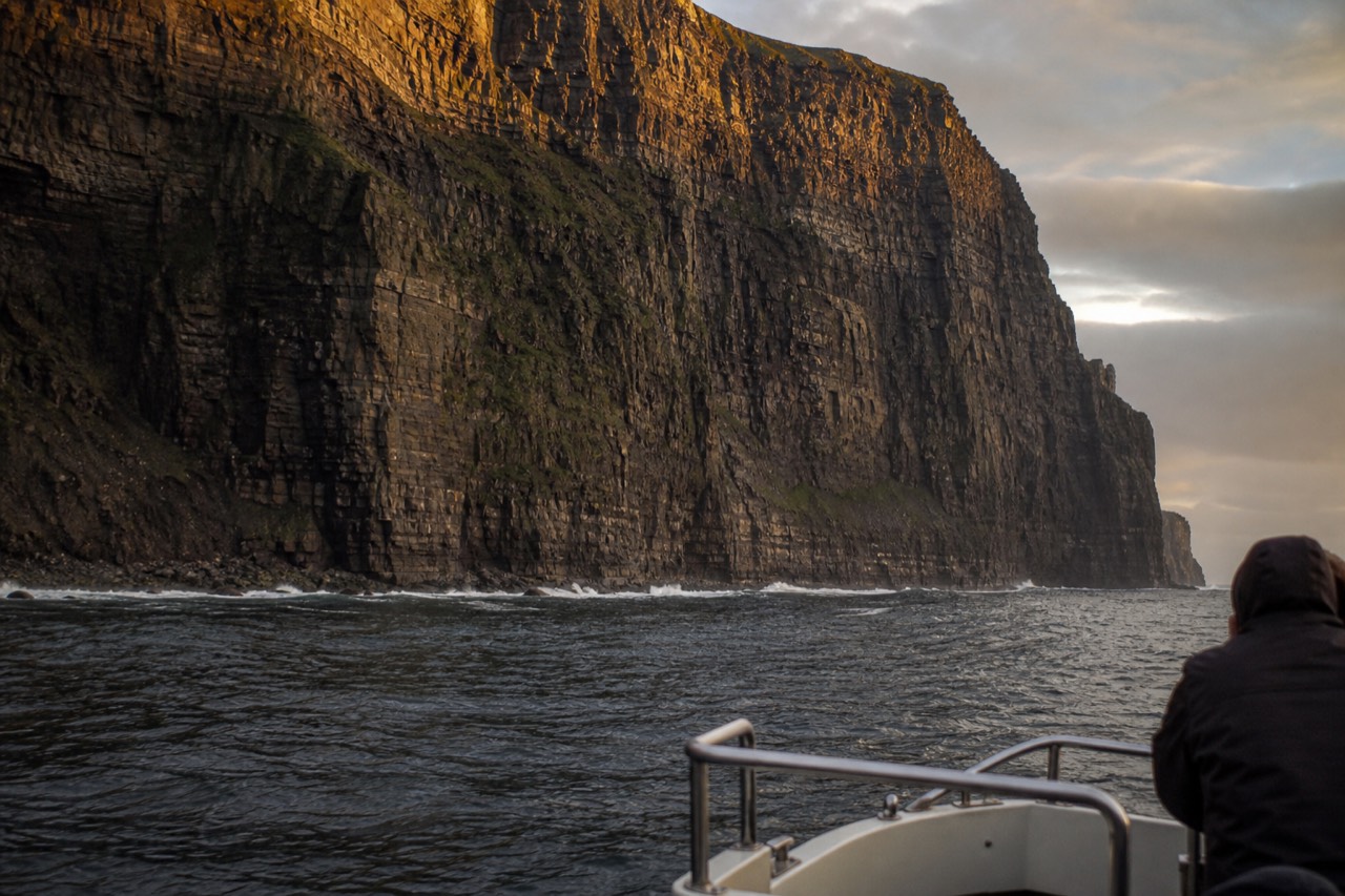 The Cliffs of Moher seen from a small boat at their base, the vast dark rock wall filling the frame above a band of white surf and grey Atlantic swell.