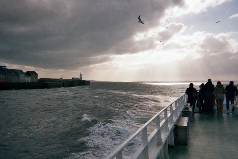 An Aran Islands ferry departing the docks at Galway City, gulls overhead and the wide expanse of Galway Bay opening out ahead under a bright sky.
