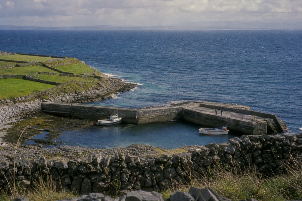 Stone-walled green fields and a small harbour on Inis Mór, the largest Aran island, with the Atlantic stretching out toward a distant hazy mainland.