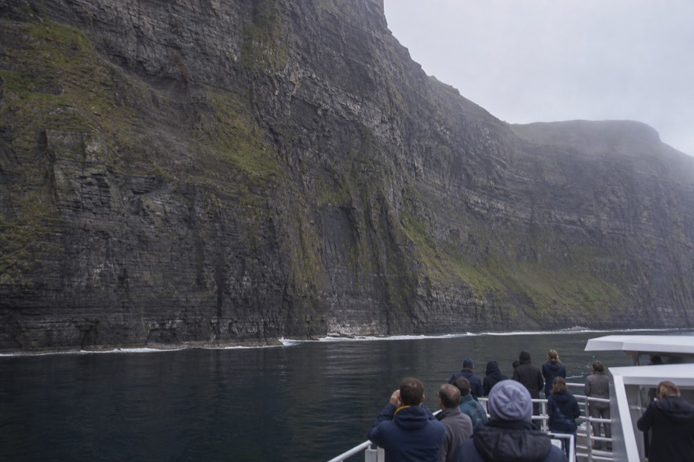 A dedicated cruise boat close beneath the Cliffs of Moher, the enormous layered rock face filling the frame above a thin line of white surf and passengers gazing upward.