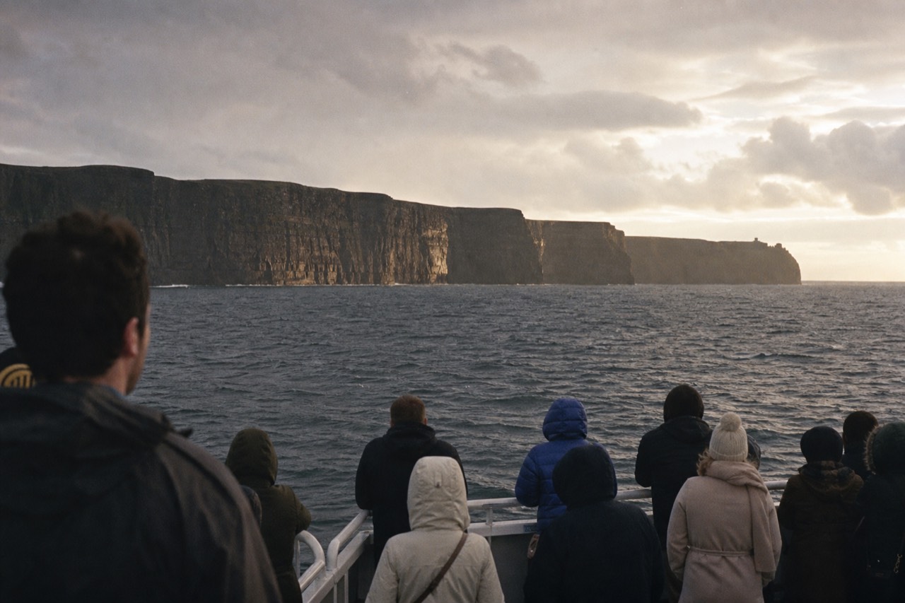 Passengers on the deck of an Aran Islands ferry looking toward the Cliffs of Moher across grey-blue water, the dark cliff wall stretching along the mainland coast in the distance.