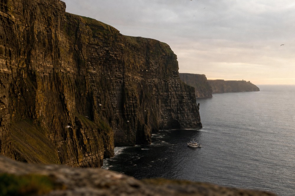 The full height of the Cliffs of Moher rising sheer from the Atlantic on a clear day, seabirds wheeling against the layered rock, a cruise boat small at the base for scale.