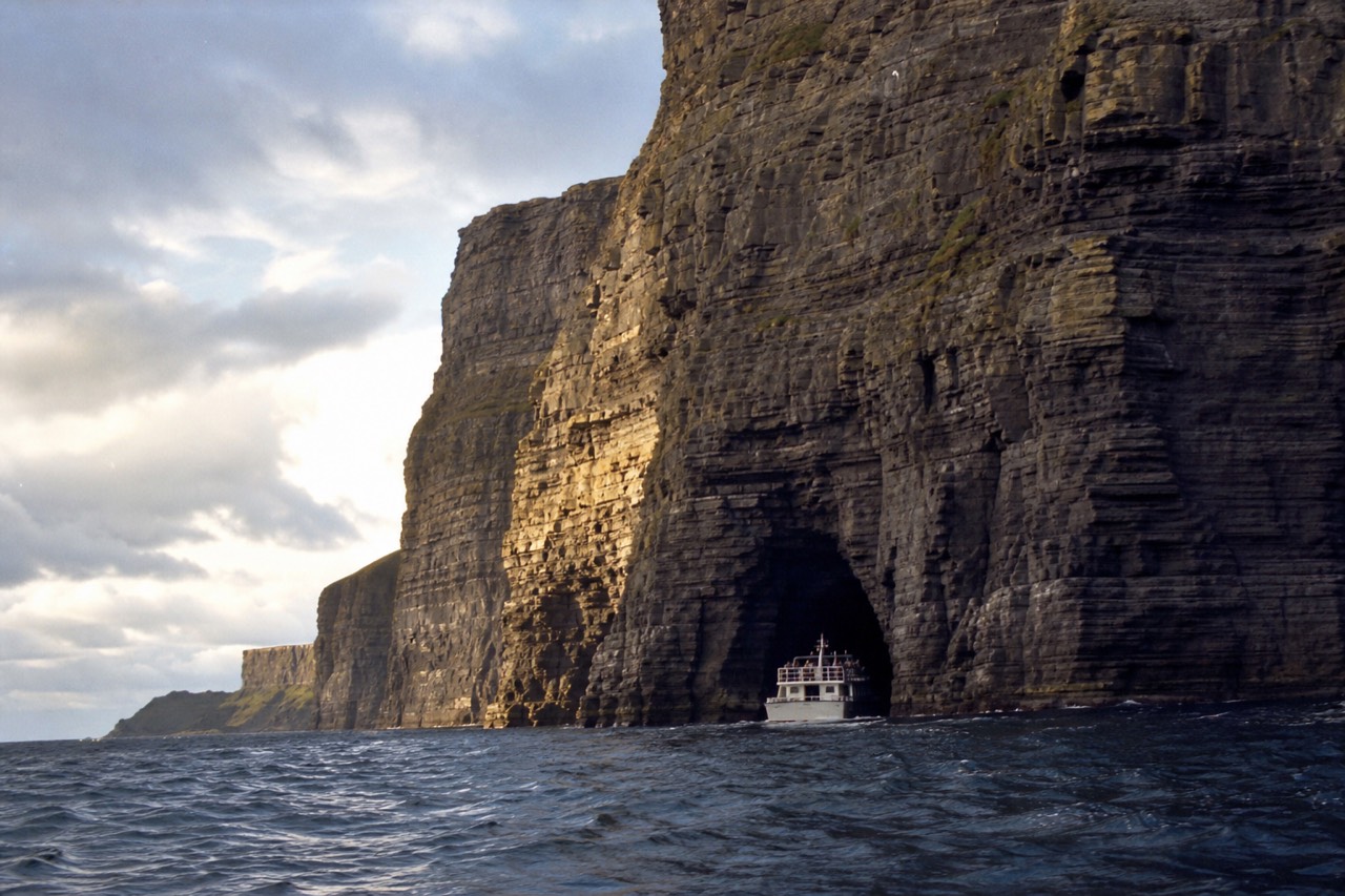 The Cliffs of Moher at golden hour from the sea, a dark sea cave cut into the base of the towering rock wall with a small cruise boat passing beneath it.
