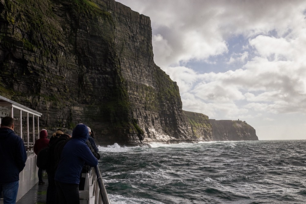 A cliff-cruise boat riding low beneath the towering layered wall of the Cliffs of Moher, passengers along the rail looking up at the rock face streaked with green.