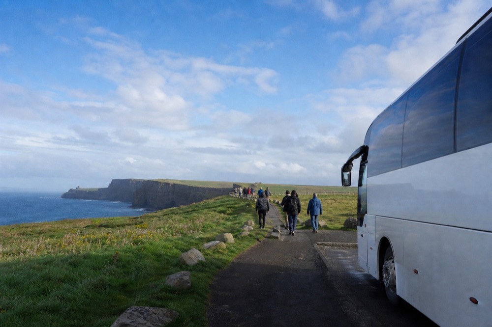 A tour coach parked at a green Irish coastal viewpoint, passengers walking a paved path toward the Cliffs of Moher clifftop under a bright, windy sky.
