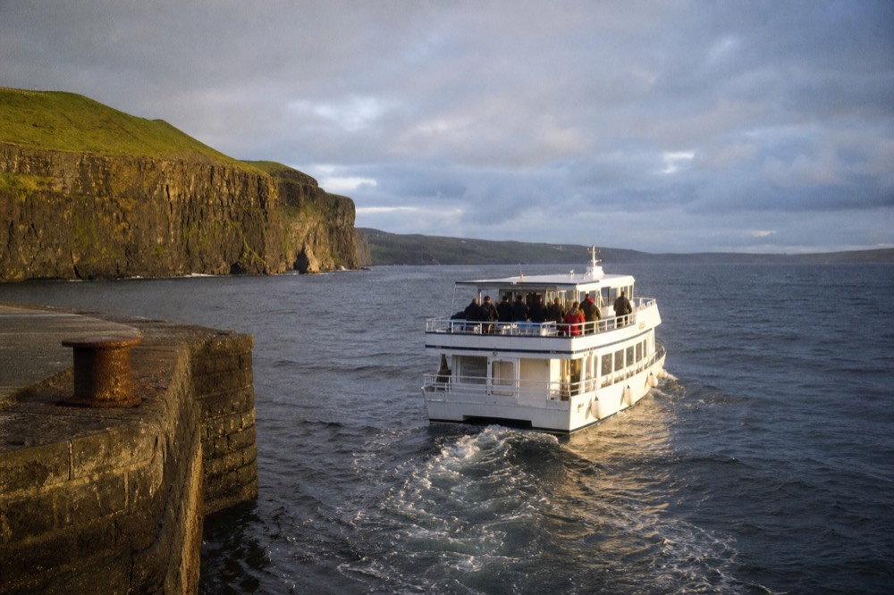 A white cliff-cruise boat pulling away from the stone jetty at Doolin Pier, green County Clare hills behind it and open grey Atlantic water ahead.