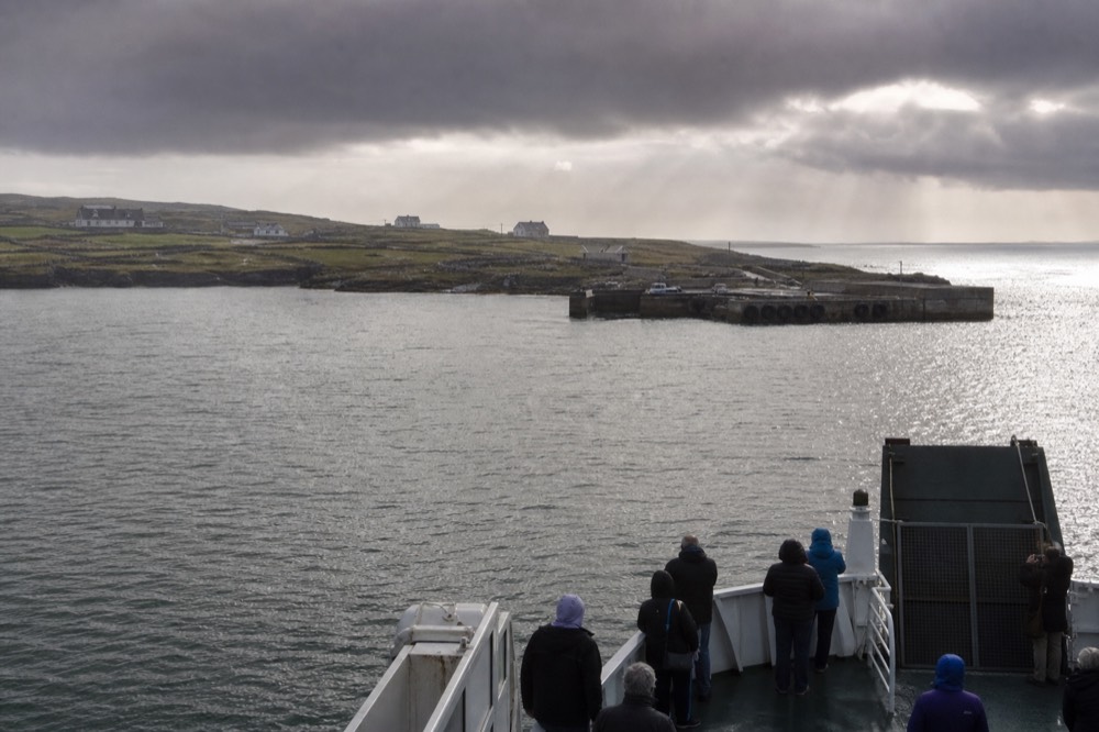 A low green Aran island edged with grey stone walls seen across calm water, a small ferry nearing its harbour with the wide Atlantic beyond.