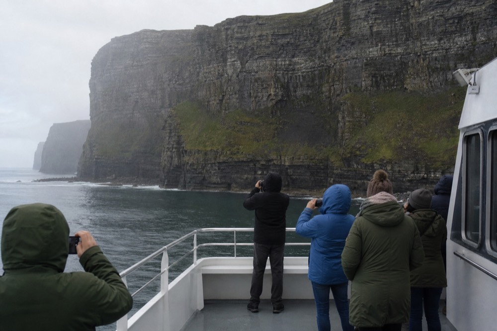 Passengers along the open deck of a cliff-cruise boat photographing the Cliffs of Moher, the layered rock wall rising close behind them over a gentle swell.