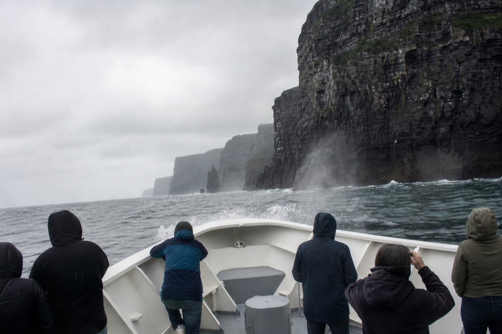 The bow of a cliff-cruise boat riding over a grey Atlantic swell at the base of the Cliffs of Moher, spray flying and the dark rock wall looming close on one side.