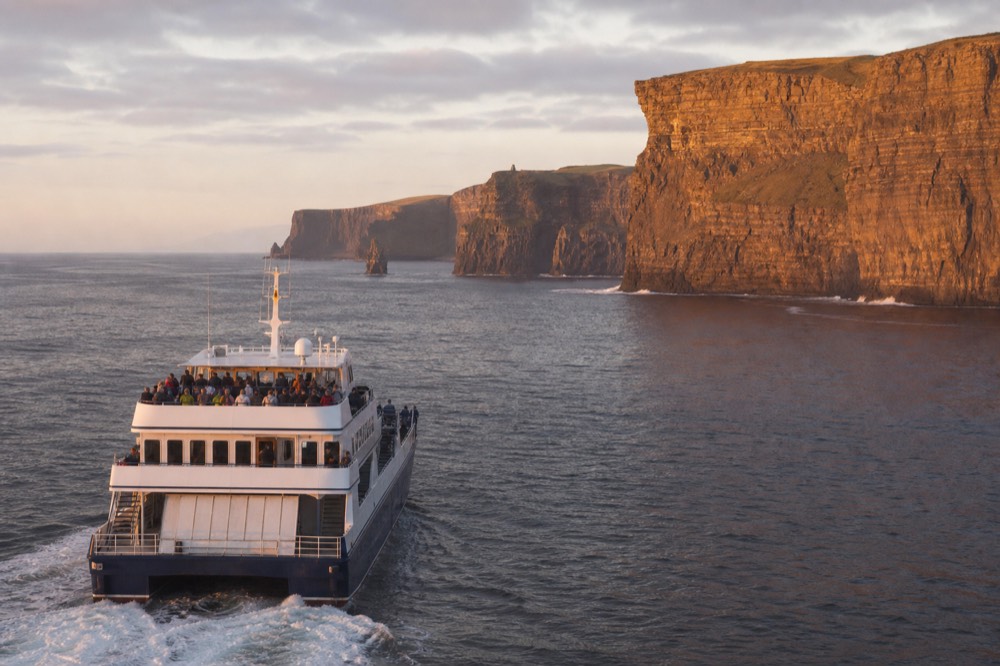 The Cliffs of Moher receding behind a returning cruise boat in soft late-afternoon light, the long rock wall glowing amber above a calm sea.