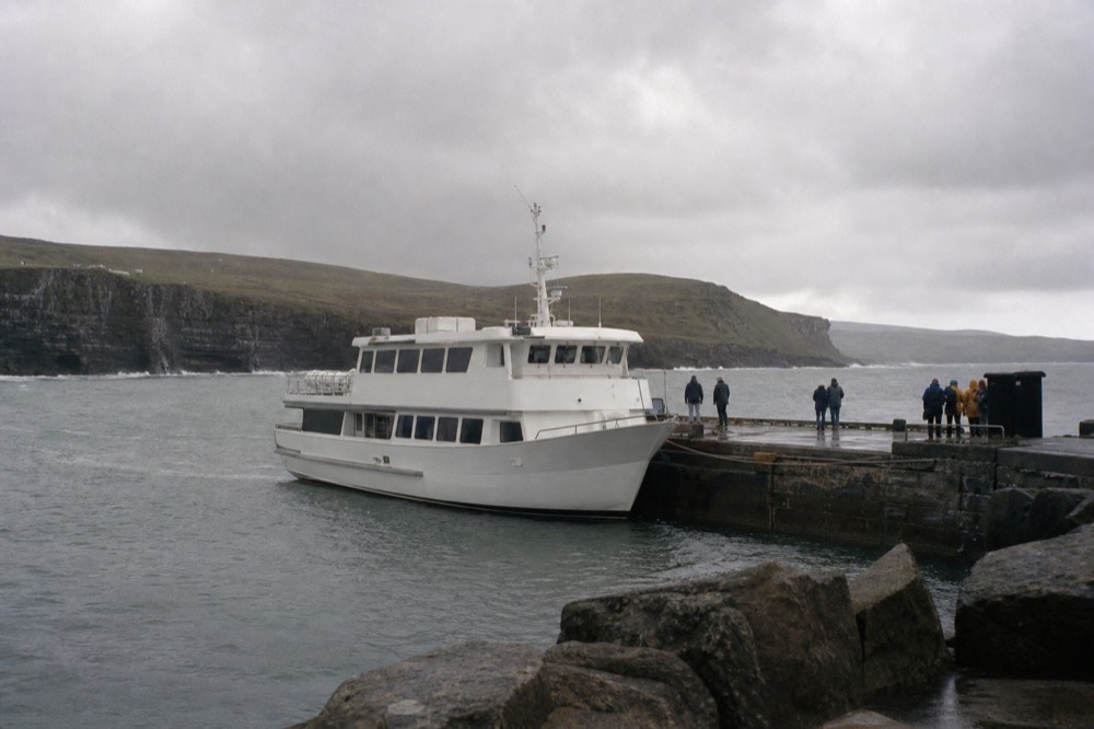 Doolin Pier in early spring, one white cruise boat moored at the stone quay under low grey cloud, the Clare hills bare and the sea a flat pewter grey.