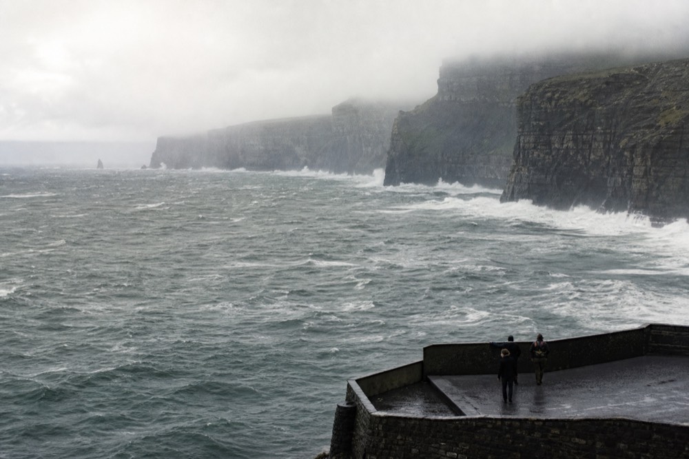 A grey Atlantic swell breaking white against the base of the Cliffs of Moher on a windy autumn day, the sea heaving and the cliff tops lost in low cloud.