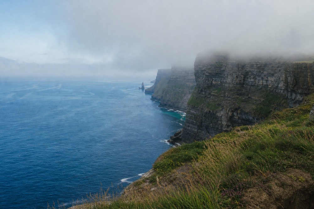 Low fog rolling across the top of the Cliffs of Moher on a still summer morning, the upper cliff hidden in grey while the sea below stays calm and pale.