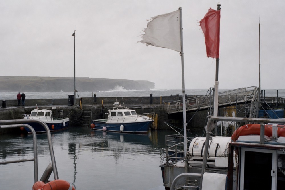 An autumn afternoon at Doolin Pier with the boats moored and a cancellation notice, wind whipping the flags and a heavy grey sea rolling beyond the harbour wall.