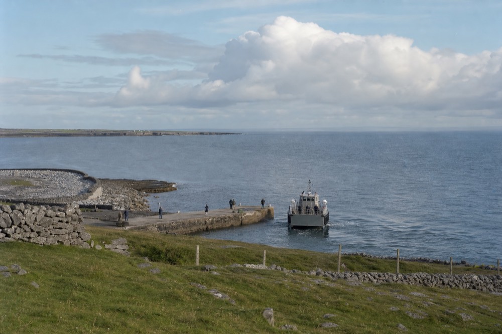 A small ferry approaching the stone harbour on Inis Oirr, low grey limestone walls and green fields behind it, the Atlantic wide and calm under a pale sky.